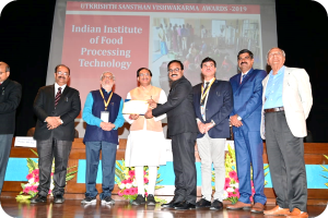 Eine Gruppe Männer steht auf einer Bühne, zwei halten eine Urkunde, mit einem Bildschirm im Hintergrund mit 'Indian Institute of Food Processing Technology Awards 2019' und Blumenschmuck.