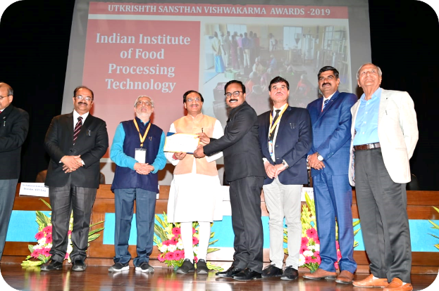 Eine Gruppe Männer steht auf einer Bühne, zwei halten eine Urkunde, mit einem Bildschirm im Hintergrund mit 'Indian Institute of Food Processing Technology Awards 2019' und Blumenschmuck.