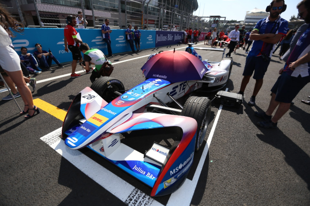 Formula E-Rennwagen mit weißer, blauer, roter und schwarzer Lackierung auf einer Rennstrecke geparkt, umgeben von Menschen, mit einem Schirm im Vordergrund und Rennstreckenabsperrungen, Gebäuden, Bäumen und einem klaren Himmel im Hintergrund.