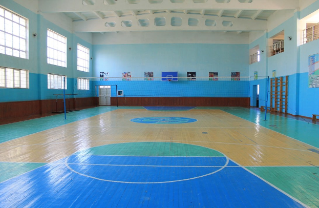 Großer Turnsaal mit blau-weißem Boden, einem Basketballfeld, Netz, Stangen, einer Tür, Fenstern, Wandplakaten, einem Geländer und einem Dach.