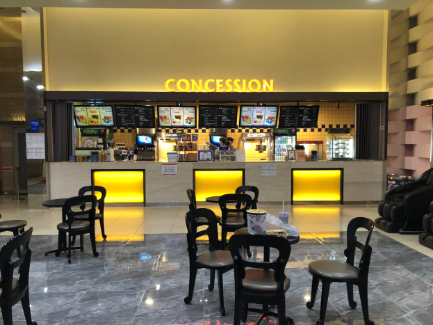 Imbissrestaurant in einem Einkaufszentrum mit Tischen, Stühlen, Gegenständen auf Tischen, einer Theke mit Personal, Texttafeln, Deckenbeleuchtung, Sofas auf der rechten Seite und Glastüren auf der linken Seite.