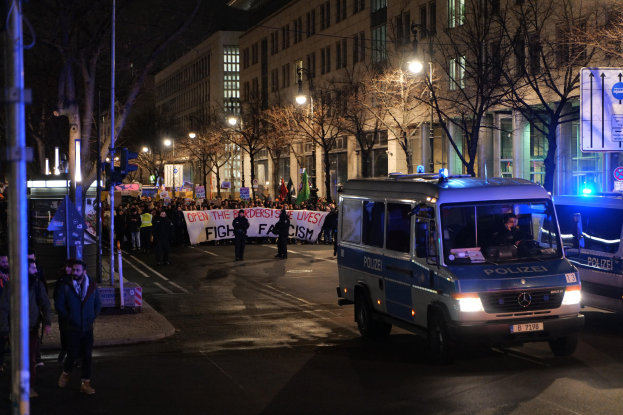 Eine Gruppe von Menschen, die nachts eine Straße entlanggehen, mit einem parkenden Polizeiwagen im Vordergrund, Bäumen und Gebäuden an der Straße und einer Banner im Hintergrund.