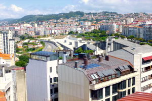 Eine Stadtansicht von einem hohen Standpunkt aus, die zahlreiche Gebäude, Bäume, Straßen, Fahrzeuge und ferne Berge unter einem bewölkten Himmel zeigt, mit Solarpanelen auf dem Dach eines Gebäudes.