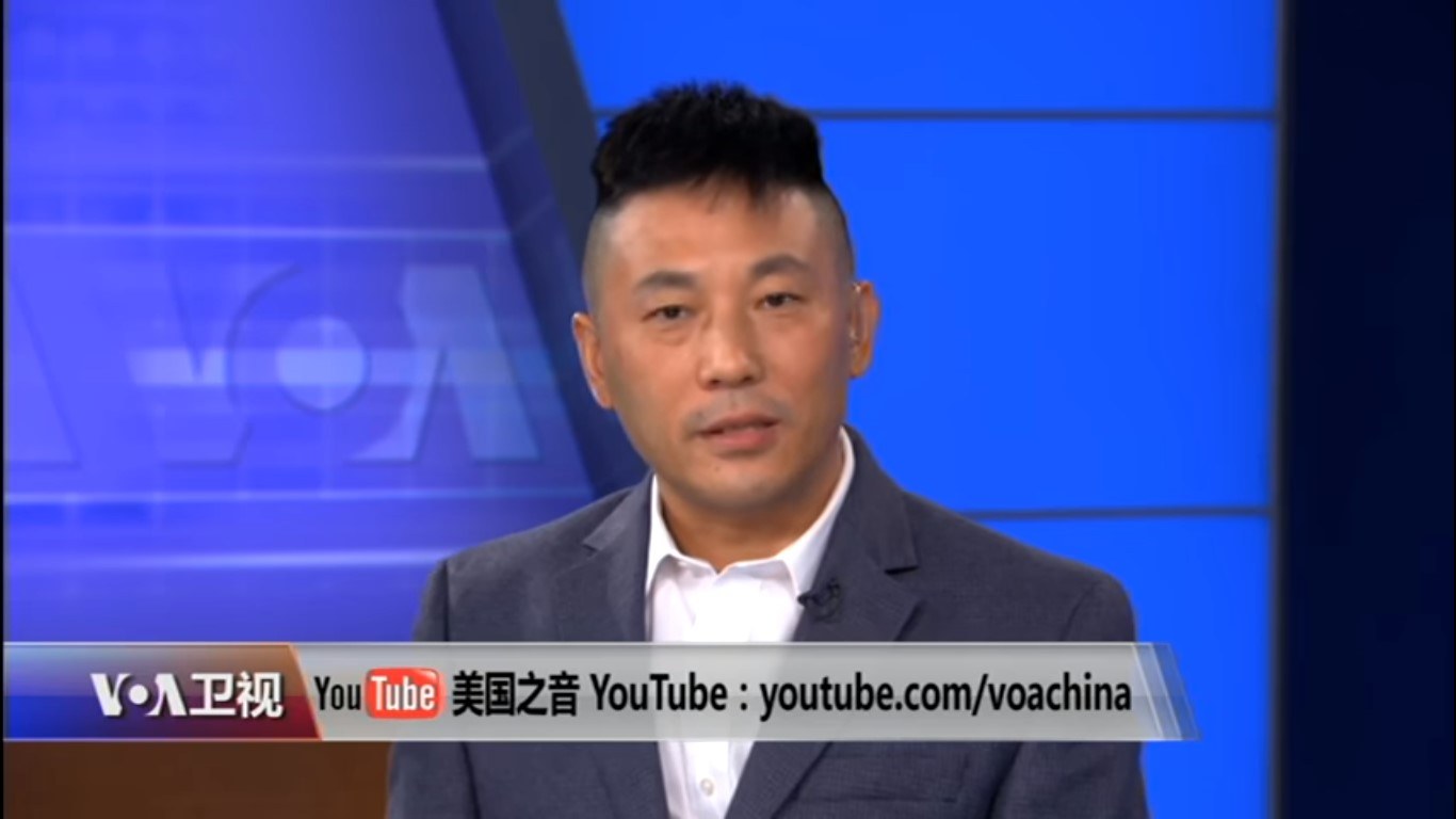 Ein chinesischer Nachrichtensprecher im Anzug und Schlips auf einem TV-Bildschirm mit blauem Hintergrund und Text am unteren Bildrand.