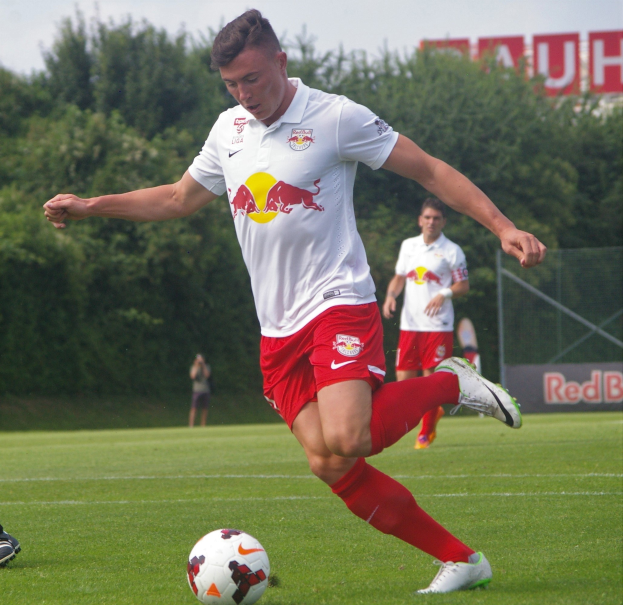 Ein professioneller Fußballspieler in einem roten Trikot schießt einen Ball auf einem Rasenfeld mit Bäumen und einem klaren blauen Himmel im Hintergrund, in der Nähe eines "RB Leipzig"-Schildes.