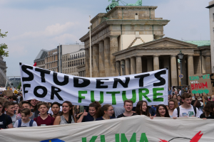 Eine Gruppe von Schülern marschiert in Berlin mit einem bunten "Students for Future"-Schild vor einer Kulisse aus Gebäuden, Bäumen und Himmel.