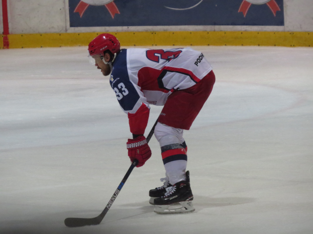 Hockey player in full gear skating on ice with a banner featuring text and logos in the background.