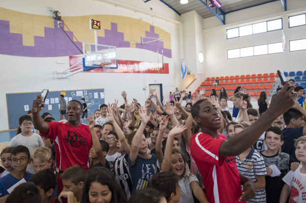 Kinder auf einem Basketballplatz mit Handys stehend, mit einer Anschlagtafel, Uhr, Torpfosten, Basketballkorb, Deckenleuchten, Stühlen und Fenstern im Hintergrund.