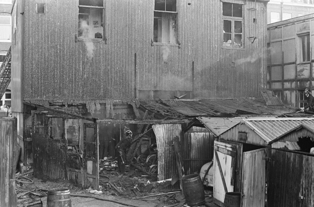 Schwarzes und weißes Foto eines feuerbeschädigten Gebäudes mit verstreuten Gegenständen wie Fässern und Holzgegenständen, einigen Menschen in der Nähe und Hintergrundgebäuden mit Fenstern, einer Leiter und einem Pfahl.