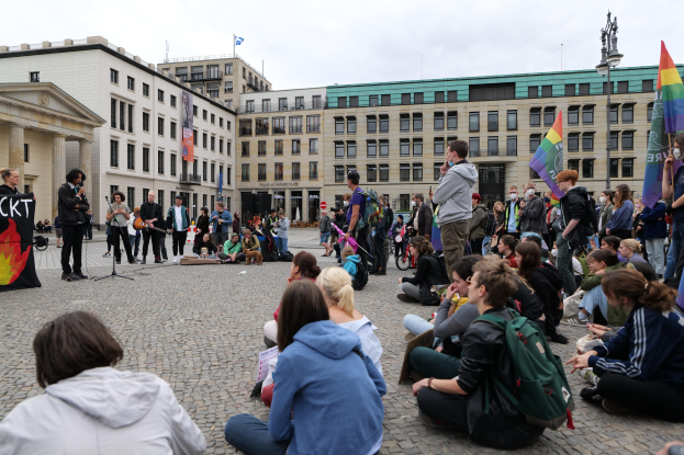 Eine Gruppe von Menschen, die auf dem Boden vor einer Menge mit Fahnen und Spruchbändern sitzen, mit einer Person, die in ein Mikrofon spricht, einer Statue und Gebäuden im Hintergrund während einer Anti-Schwulen-Demo in Berlin.