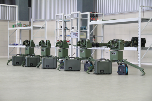 Eine Gruppe von Milit├Ąrrobotern, die auf einem Boden mit Koffern und anderen Gegenst├Ąnden auf umliegenden Gestellen positioniert sind, vor einer Wand mit einem Gel├Ąnder im Hintergrund.