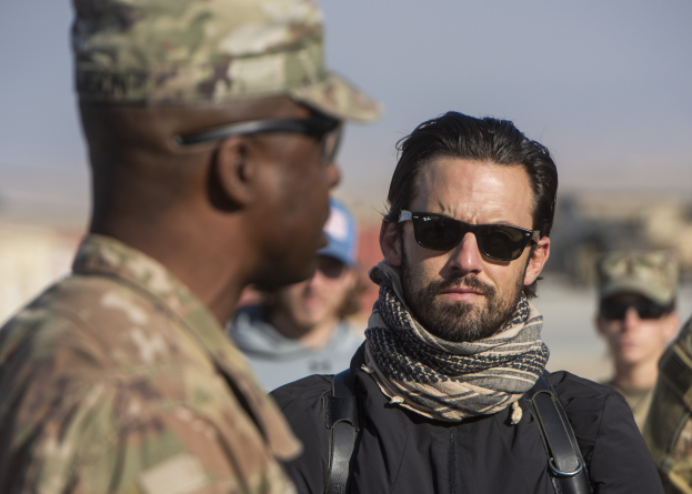 Zwei Männer in einem Gespräch, einer in militärischer Uniform und der andere mit einem Schal und einer Schutzbrille, mit einem unscharfen Hintergrund.