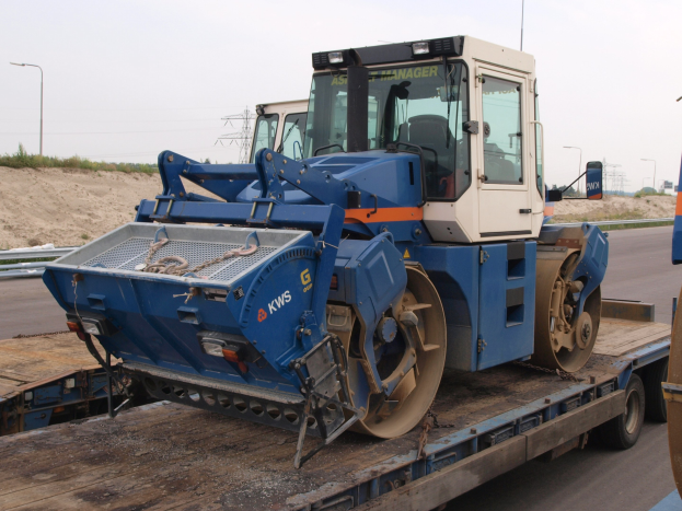 Ein Walzenstapler wird auf einen Tieflader geladen, im Hintergrund sind Laternenpfähle, Bäume und ein klarer blauer Himmel zu sehen.