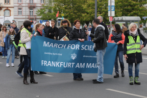 Eine bunte Gruppe von Menschen marschiert eine Straße entlang und hält ein "March for Science Frankfurt am Main"-Schild hoch, mit Bäumen, Gebäuden und einem klaren blauen Himmel im Hintergrund.