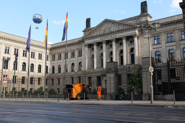 Großes modernes Gebäude mit vielen Fenstern, als Bundestag in Berlin, Deutschland identifiziert, umgeben von Straßeninfrastruktur, Fußgängern und Flaggen, unter einem bewölkten Himmel.