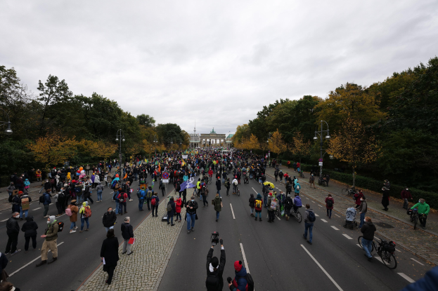 Eine große Gruppe von Menschen marschiert auf einer baumbestandenen Straße in Berlin, einige halten Kameras, mit einem Gebäude und einem klaren Himmel im Hintergrund.