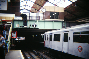 Ein Zug fährt auf einem Bahnsteig ein, wo Menschen warten, mit einer Ampel, einem Schild, einer Uhr, Metallstangen und einer Decke mit Deckenleuchten.