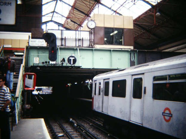 Ein Zug fährt auf einem Bahnsteig ein, wo Menschen warten, mit einer Ampel, einem Schild, einer Uhr, Metallstangen und einer Decke mit Deckenleuchten.