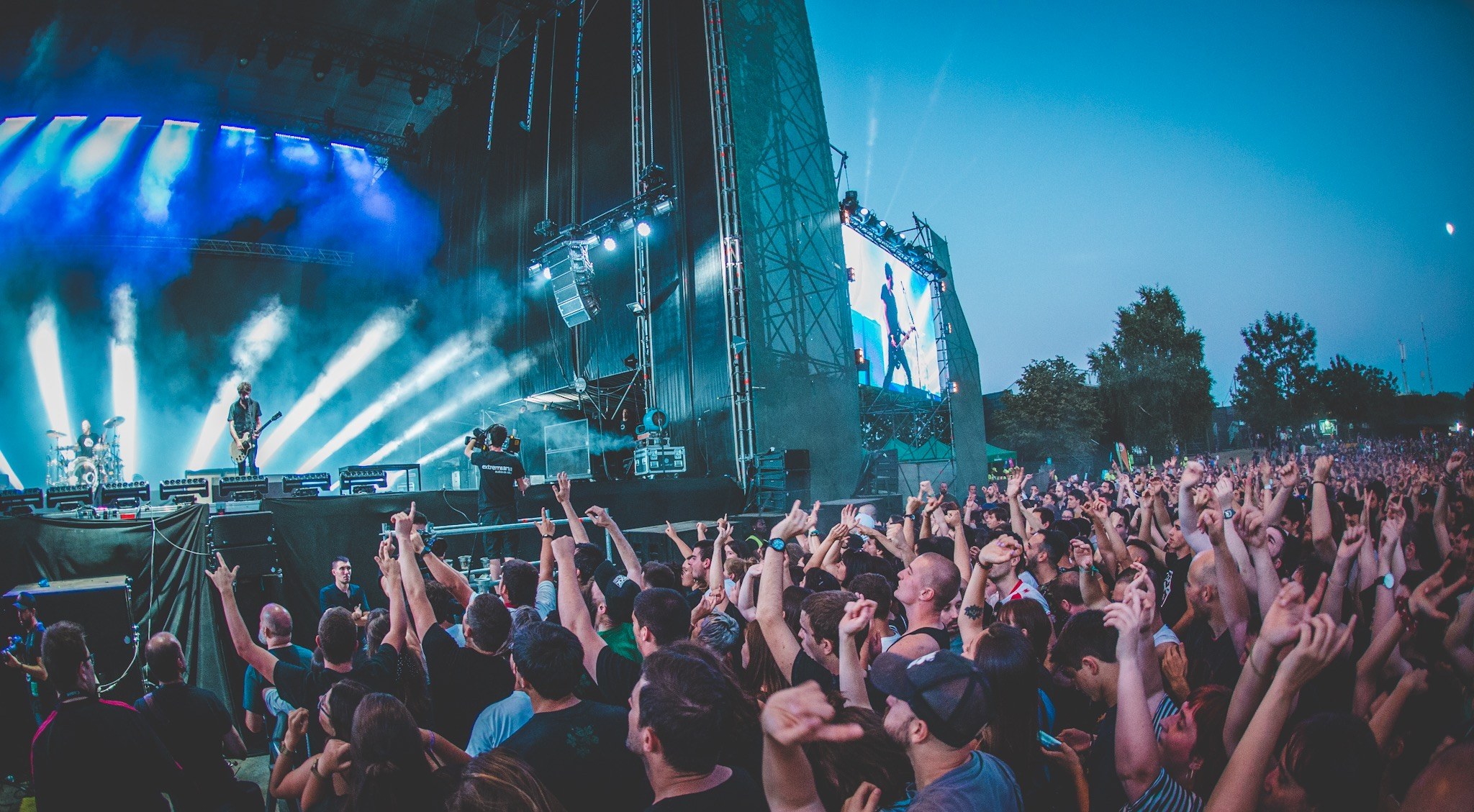 Menschenmenge auf einem Musikfestival mit erhobenen Händen vor einer Bühne mit Instrumenten, Lichtern und einem Bildschirm, vor Bäumen und einem klaren blauen Himmel.
