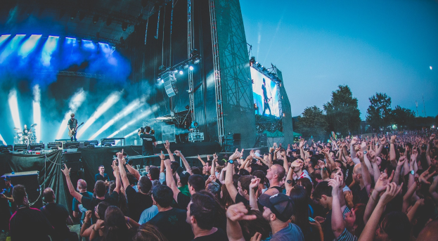 Menschenmenge auf einem Musikfestival mit erhobenen Händen vor einer Bühne mit Instrumenten, Lichtern und einem Bildschirm, vor Bäumen und einem klaren blauen Himmel.