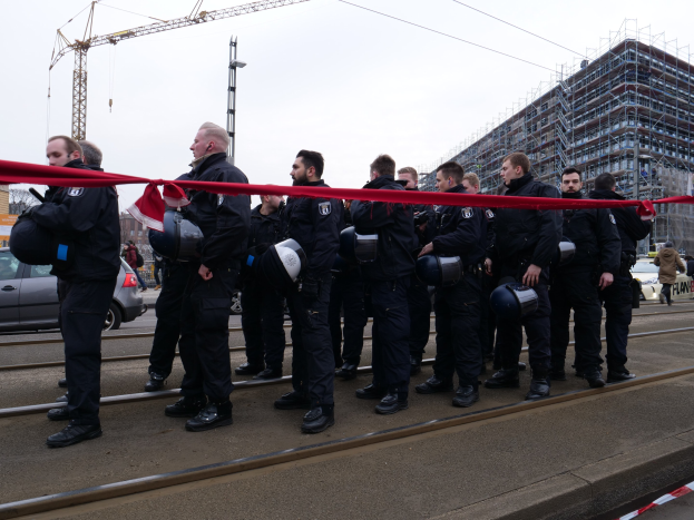 Eine Gruppe von Polizisten in schwarzen Uniformen und Helmen steht in einer Reihe auf einem Bahngleis, mit einem roten Band davor, vor einer Kulisse aus Fahrzeugen, Passanten, Gebäuden und einem klaren blauen Himmel.