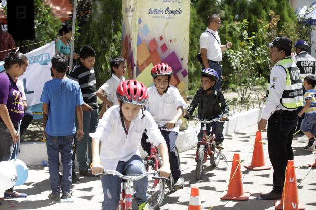 Eine Gruppe von Kindern, die Fahrräder auf einer Straße mit Verkehrskegeln fahren, einige tragen Helme, andere stehen daneben, mit einer Fahne, Bäumen und Gebäuden im Hintergrund.