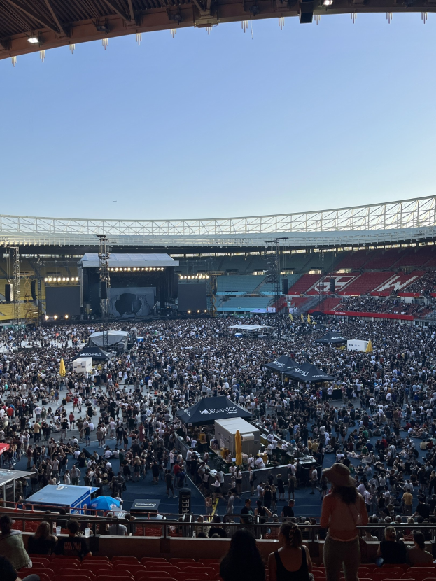 Eine große Menschenmenge, die auf Sitzplätzen und Stehplätzen in einer Arena mit Zelten, Geländern und Scheinwerfern sitzt, vor einem Himmel als Hintergrund, was auf ein Konzert in der O2 Arena in London hindeutet.