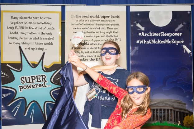 Zwei junge Mädchen in Superheldenkostümen posieren für ein Foto vor einem blauen Vorhang bei einem Schulereignis, eine hält einen Stoff in der Hand, während beide Masken tragen.