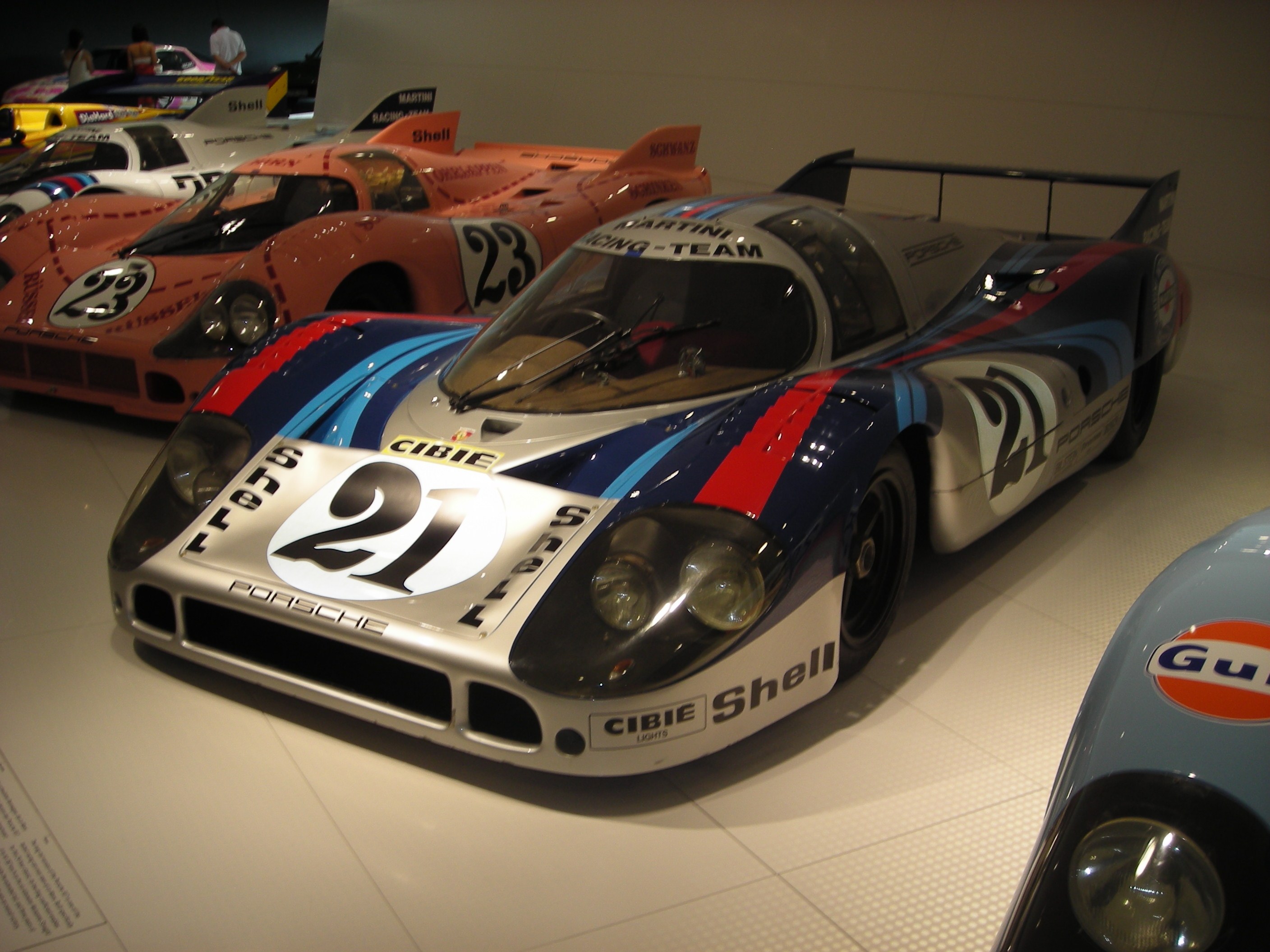 Ein Porsche 917K Rennwagen auf einer Museumsausstellung, umgeben von anderen Sportwagen, mit Menschen in der Nähe und einer Wand im Hintergrund.