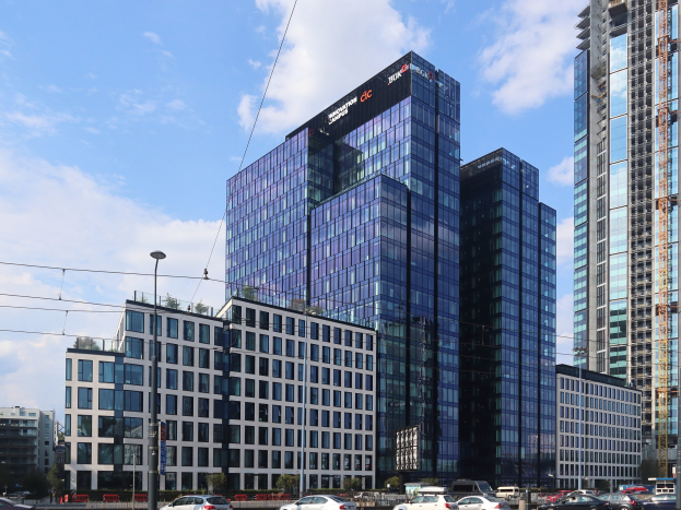 Zentrale der Deutschen Bank in Frankfurt, Deutschland, ein großes mehrfenstriges Gebäude umgeben von städtischer Infrastruktur einschließlich Straßenlaternen, Fahrzeugen und Grünflächen unter einem bewölkten Himmel.