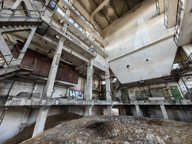 Verlassene Fabrikhalle mit einem großen Haufen Erde im Vordergrund, mit Treppen, Geländern, Säulen, Wänden, Fenstern, Lampen und einem Sandhaufen unten.