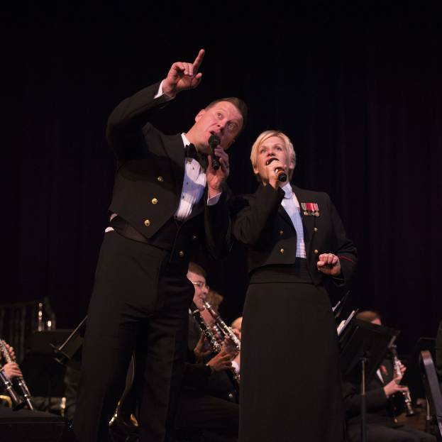 Ein Mann und eine Frau in Smokings stehen auf einer Bühne und halten Mikrofone, mit einem dunklen Hintergrund und einem sitzenden Orchester dahinter.