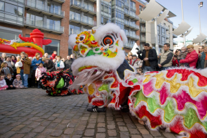 Ein lebendiges chinesisches Neujahrsfest in Amsterdam mit einer Löwen-Tanz-Performance und einer Menge Schaulustiger, einige mit Kameras, vor einem Hintergrund aus Gebäuden, Laternenmasten und einem klaren blauen Himmel.