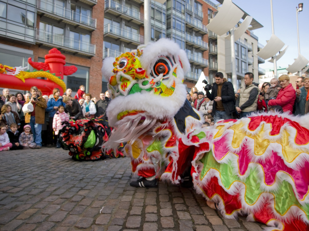 Ein lebendiges chinesisches Neujahrsfest in Amsterdam mit einer Löwen-Tanz-Performance und einer Menge Schaulustiger, einige mit Kameras, vor einem Hintergrund aus Gebäuden, Laternenmasten und einem klaren blauen Himmel.