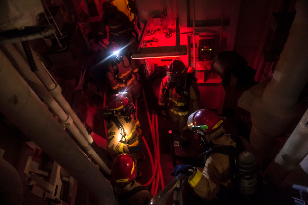 Feuerwehrmänner in Helmen und Sauerstoffflaschen stehen in einem dunklen Raum mit Rohren auf beiden Seiten, mit einer Maschine und anderen Gegenständen im Hintergrund, was auf einen Rettungseinsatz hinweist.