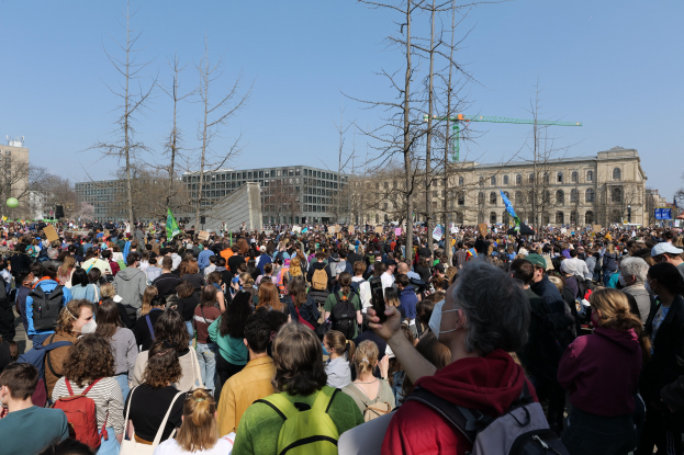 Große Menge Menschen mit Schildern und Taschen vor einem Gebäude mit Bäumen und einem klaren Himmel, was auf eine Klimaprotestveranstaltung hinweist.