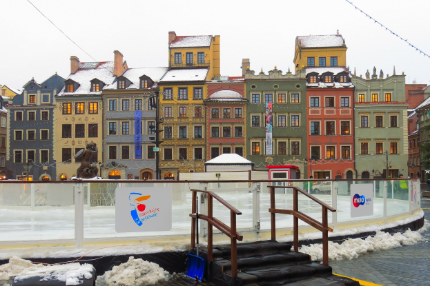 Eislaufbahn in einem Stadtplatz umgeben von Gebäuden, Treppen, Geländern, Informationsschildern, Laternen, Bäumen und schneebedeckter Erde unter einem Himmel.