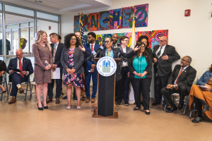 Eine Gruppe von Menschen vor einem Podium mit einem Mikrofon und einer Tafel, auf der 'Sancho City Councilman Joins City Council' steht, einige sitzen, andere stehen, Flaggen, eine Tafel, ein Feuerlöscher und Deckenleuchten im Hintergrund.