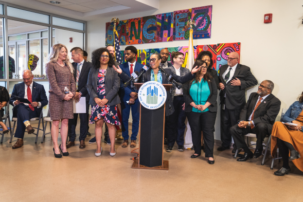 Eine Gruppe von Menschen vor einem Podium mit einem Mikrofon und einer Tafel, auf der 'Sancho City Councilman Joins City Council' steht, einige sitzen, andere stehen, Flaggen, eine Tafel, ein Feuerlöscher und Deckenleuchten im Hintergrund.