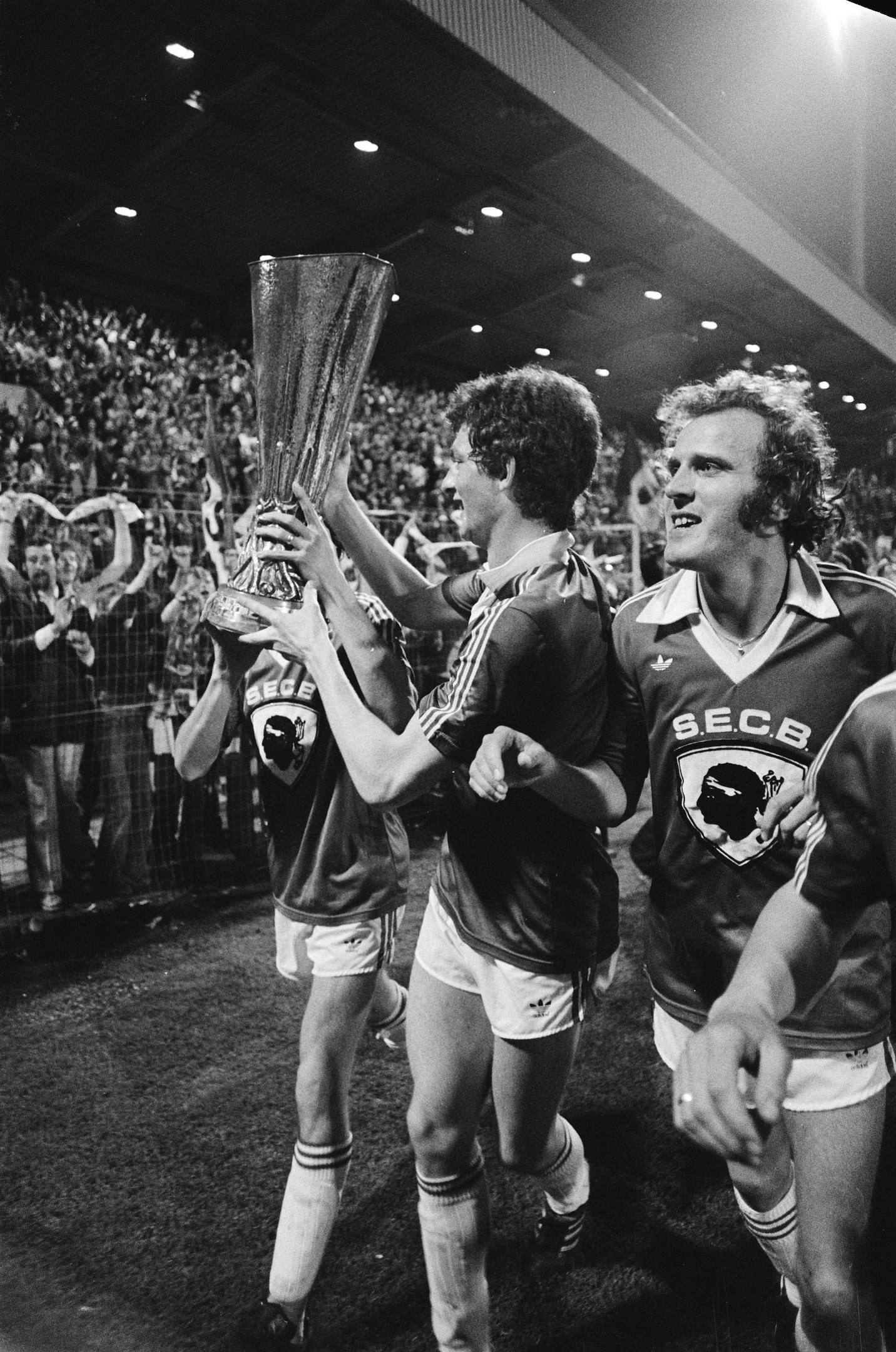 Schwarzes und weißes Bild von Fußballspielern, die mit einem Pokal auf einem Feld feiern und von Zuschauern hinter einem Zaun und überdachten Stadionlampen umgeben sind.