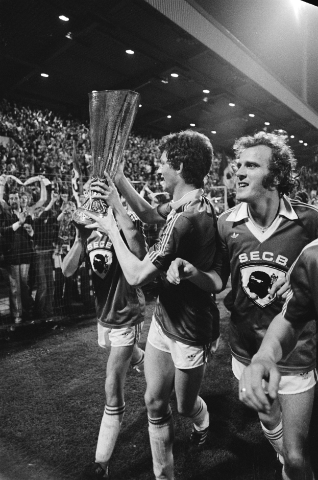 Schwarzes und weißes Bild von Fußballspielern, die mit einem Pokal auf einem Feld feiern und von Zuschauern hinter einem Zaun und überdachten Stadionlampen umgeben sind.