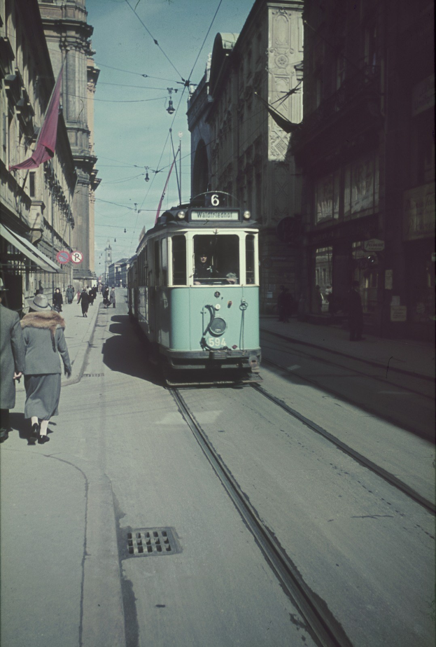 Altes Schwarz-Weiß-Foto einer Straßenbahn, die auf einer Stadtstraße mit Fußgängern auf dem linken Gehweg fährt, mit Gebäuden und Strommasten im Hintergrund unter einem klaren Himmel.
