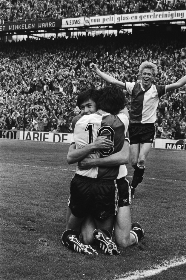 Drei Fußballspieler feiern auf dem Boden nach einem Tor, mit jubelnder Menge und schwenkenden Fahnen in einem schwarz-weißen Stadion.