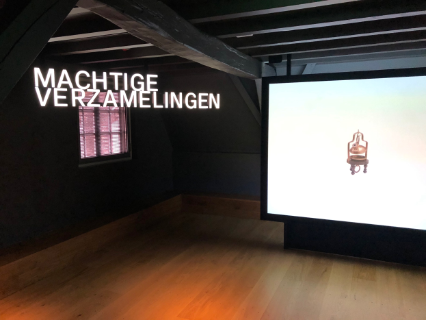 Installationsansicht der Ausstellung 'Machtige Sammlung' im Museum of Modern Art in Berlin, mit einem großen Bildschirm mit Wandtext, einem Fenster links und sichtbarem Dach.