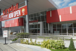 Stadtmalleingangsschild mit der Aufschrift "Stadt Mall Now Open" mit umgebender Vegetation, einem Metallzaun, Menschen, einem Auto, Pfosten, einer Fahne, Lichtern und einem bewölkten Himmel.