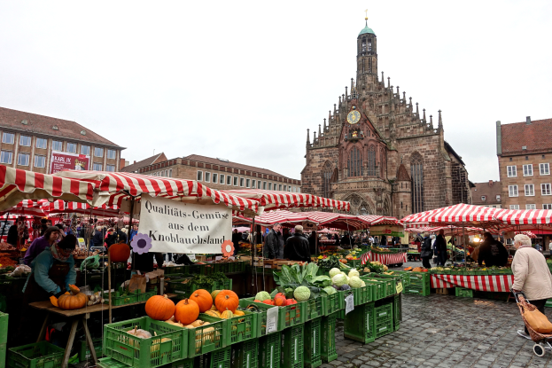 Ein belebter Markt in Nürnberg, Deutschland, mit verschiedenen Obst- und Gemüsesorten, Menschen, die umherlaufen, Zelten, Gebäuden mit Fenstern und einem Kirchturm im Hintergrund unter einem sichtbaren Himmel.