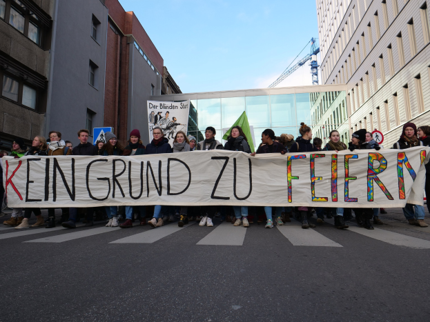 Gruppe von Menschen mit einem Banner "Kein Grund zu Feiern" im Protest gegen Sparmaßnahmen der deutschen Regierung, mit Gebäuden, einem Schild, einem weiteren Textbanner, einem Kran und einem bewölkten Himmel im Hintergrund.