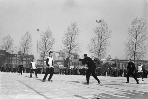 Eine Gruppe von Menschen, die im Schnee Eishockey spielen, mit Bäumen, Gebäuden mit Fenstern, Laternen und einem klaren Himmel im Hintergrund, dargestellt in Schwarz-Weiß.