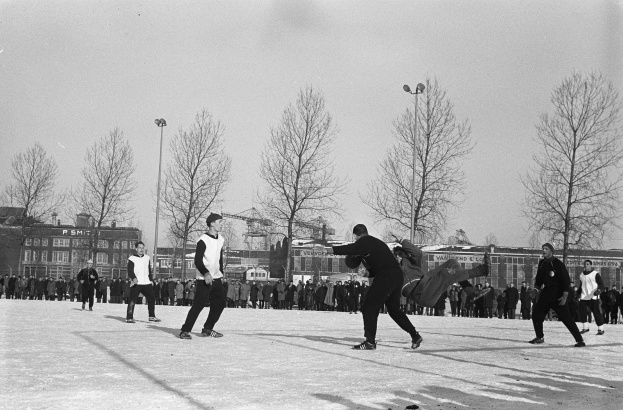 Eine Gruppe von Menschen, die im Schnee Eishockey spielen, mit Bäumen, Gebäuden mit Fenstern, Laternen und einem klaren Himmel im Hintergrund, dargestellt in Schwarz-Weiß.