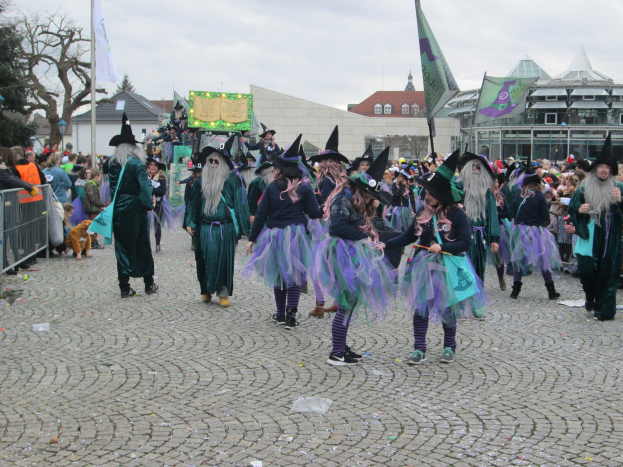 Eine Gruppe von Menschen in Hexenkostümen, die schwarze und lila Kleider und Hüte tragen und die eine Gasse aus Kopfsteinpflaster mit Flaggen, einem Geländer auf der linken Seite und Gebäuden, Bäumen und einem klaren blauen Himmel im Hintergrund entlanggehen.